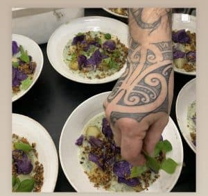 Chef with tattooed hand garnishing gourmet dish with herbs and purple chips