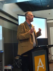 Man in brown blazer giving presentation at podium with screen behind.