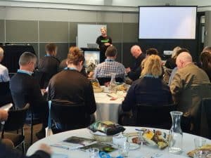 Business conference with presenter and attentive audience seated round tables.