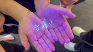 Hands under ultraviolet light showing glowing powder particles.