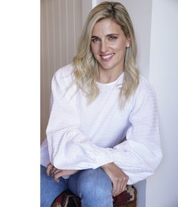 Smiling blonde woman in white blouse and blue jeans sitting.