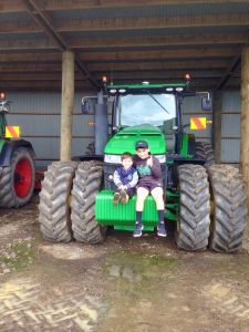 Two boys sitting on front of large green tractor in barn.
