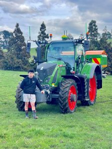 Young man standing beside green tractor on grassy field with trees.