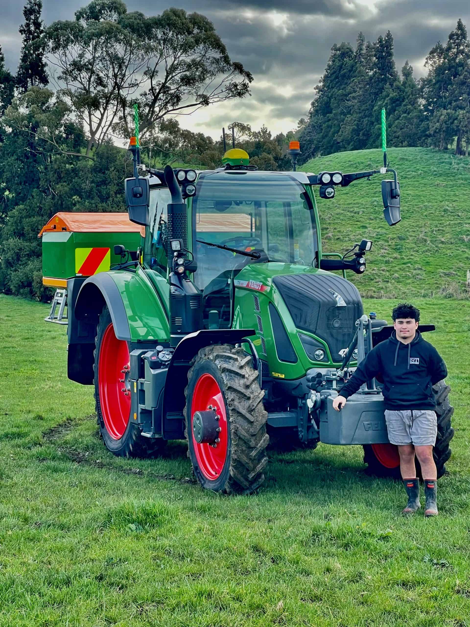 Young man standing beside green tractor on grassy field with cloudy sky